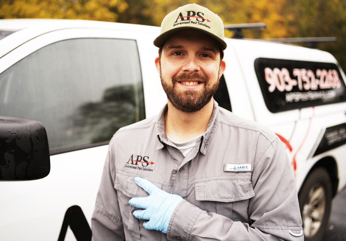A friendly APS technician standing in front of his service truck, smiling and pointing at the APS logo on his work shirt. East Texas Pest Control Service.